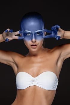 Portrait of a young woman who is posing covered with blue paint in the studio on a black background. girl&#39 s hands around the face and painted blue