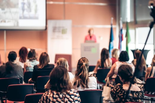 Business and entrepreneurship symposium. Female speaker giving a talk at business meeting. Audience in conference hall. Rear view of unrecognized participant in audience.