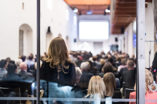 Speaker Giving a Talk at Business Meeting. Audience in the conference hall. Business and Entrepreneurship. Shot trough glass. Focus on unrecognizable woman in the audience.