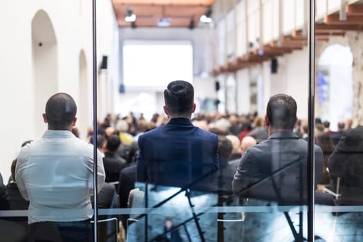 Speaker Giving a Talk at Business Meeting. Audience in the conference hall. Business and Entrepreneurship. Shot trough glass. Focus on unrecognizable man in the audience.
