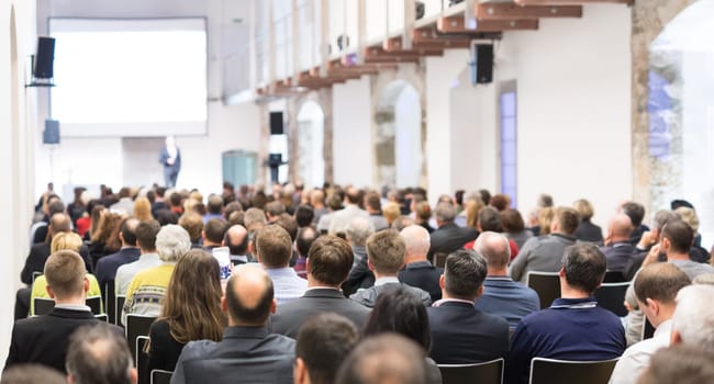 Speaker giving a talk in conference hall at business event. Audience at the conference hall. Business and Entrepreneurship concept. Focus on unrecognizable businessman in the audience.