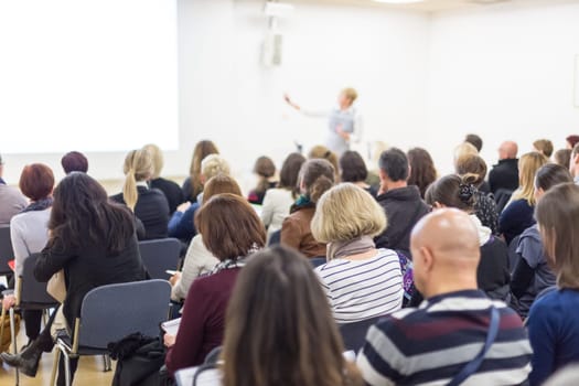 Business and entrepreneurship symposium. Female speaker giving a talk at business meeting. Audience in conference hall. Rear view of unrecognized participant in audience. Copy space on whitescreen.