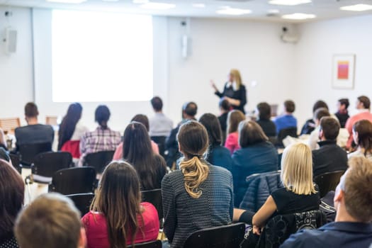 Business and entrepreneurship symposium. Female speaker giving a talk at business meeting. Audience in conference hall. Rear view of unrecognized participant in audience. Copy space on whitescreen.