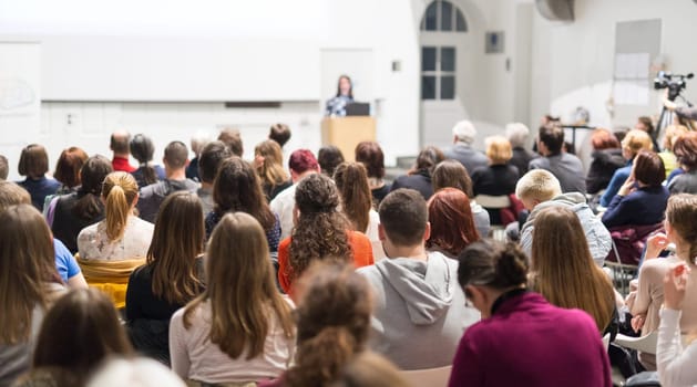 Female speaker giving presentation in lecture hall at university workshop. Audience in conference hall. Rear view of unrecognized participant in audience. Scientific conference event.