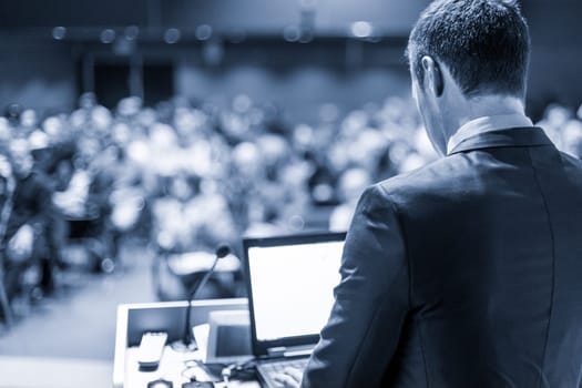 Speaker giving a talk on corporate business conference. Unrecognizable people in audience at conference hall. Business and Entrepreneurship event. Blue toned grayscale image.