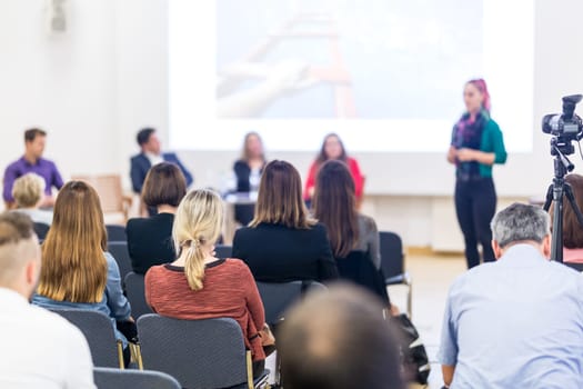 Business and entrepreneurship symposium. Female speaker giving a talk at business meeting. Audience in conference hall. Rear view of unrecognized participant in audience. Copy space on whitescreen.
