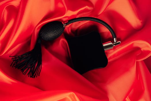 Black bottle of eau de toilette or perfume with long tassel spray pomp lies on red background of flowing fabric