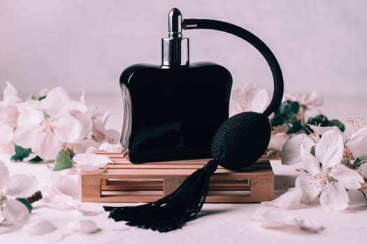 Black bottle eau de toilette or perfume with long tassel spray pomp, on wooden podium in form of pallet on light plaster surface with apple flowers