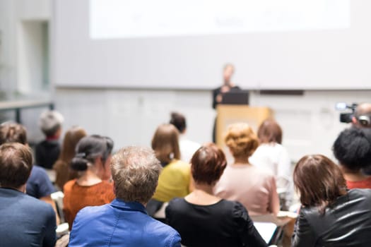 Business and entrepreneurship symposium. Female speaker giving a talk at business meeting. Audience in conference hall. Rear view of unrecognized participant in audience. Copy space on whitescreen.