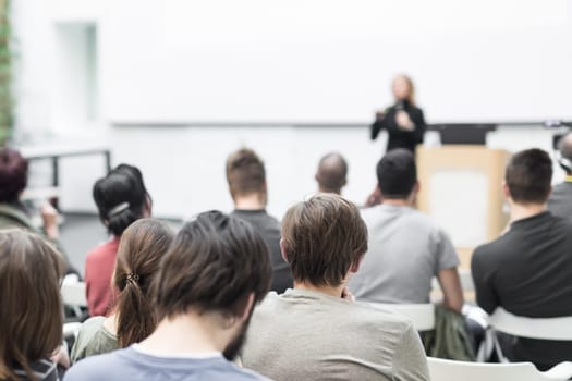 Business and entrepreneurship symposium. Female speaker giving a talk at business meeting. Audience in conference hall. Rear view of unrecognized participant in audience. Copy space on whitescreen.