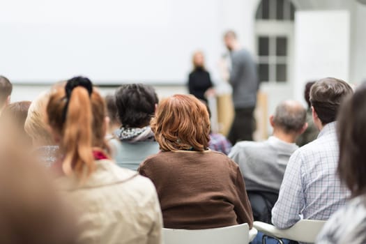 Business and entrepreneurship symposium. Female speaker giving a talk at business meeting. Audience in conference hall. Rear view of unrecognized participant in audience. Copy space on whitescreen.