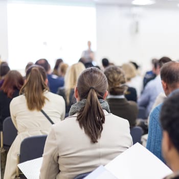 Business and entrepreneurship symposium. Female speaker giving a talk at business meeting. Audience in conference hall. Rear view of unrecognized participant in audience. Copy space on whitescreen.