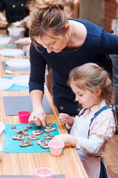 Little girl with her mom's help decorating baked cookies with colorful sprinkle and icing sugar. Kid taking part in baking workshop. Baking classes for children, aspiring little chefs. Learning to cook. Combining and stirring prepared ingredients. Real people, authentic situations