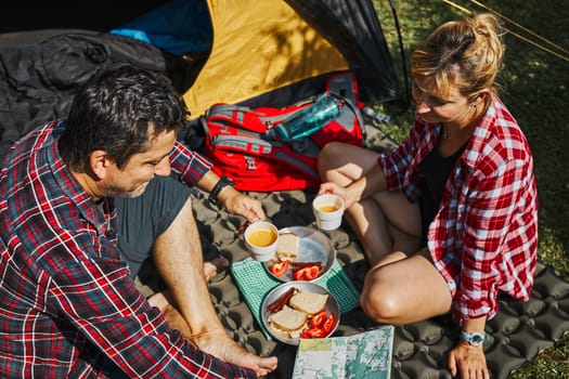 Couple having breakfast and planning their day sitting by tent at camping. People actively spending summer vacations close to nature outdoors. Concept of camp life