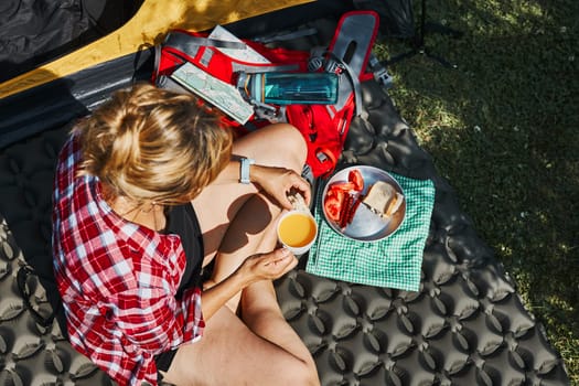 Woman having breakfast sitting by tent at camping. Female actively spending summer vacations close to nature outdoors. Concept of camp life