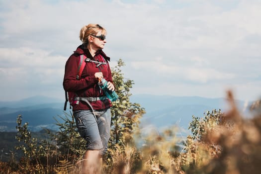 Woman with backpack hiking in mountains, spending summer vacation close to nature. Woman walking on top of hill admiring mountain landscape panorama holding bottle with water
