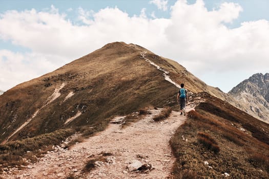 Young man with backpack hiking in a mountains, actively spending summer vacation. Rear view of teenager walking up on a hill along mountain path
