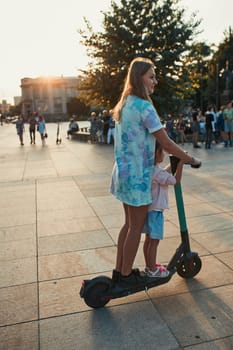 Teenager girl and her sister preschooler riding an electric scooter in the city center. Concept of happy family. Candid people, real moments, authentic situation