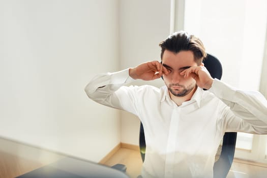 Tired exhausted young man rubbing eyes feeling eyestrain from long computer work. Man wearing white shirt sitting at computer screen