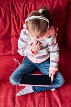 Little girl preschooler learning online solving puzzles playing educational games listetning to music and sounds using tablet and headpphones sitting on sofa at home