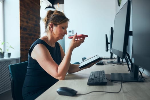 Businesswoman having business call working on tablet in office. Mature busy woman using touch pad computer standing by window in modern interior. Female manager focused on work holding digital device. Using technology