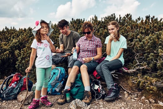 Family having break and eating snacks during trip in mountains. Family actively spending summer vacation together. Mother with her children teenagers and child sitting on trunk by mugo pine resting on peak of a mountain