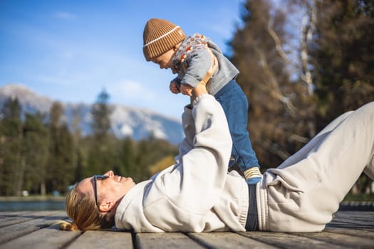 Happy family. Young mother playing with her baby boy infant oudoors on sunny autumn day. Portrait of mom and little son on wooden platform by lake. Positive human emotions, feelings, joy