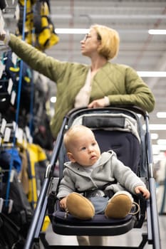 Casualy dressed mother choosing sporty shoes and clothes products in sports department of supermarket store with her infant baby boy child in stroller