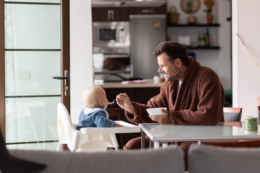 Father wearing bathrope spoon feeding hir infant baby boy child sitting in high chair at the dining table in kitchen at home in the morning