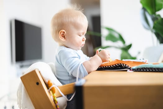 Happy infant sitting at dining table and playing with his toy in traditional scandinavian designer wooden high chair in modern bright atic home. Cute baby playing with toys.