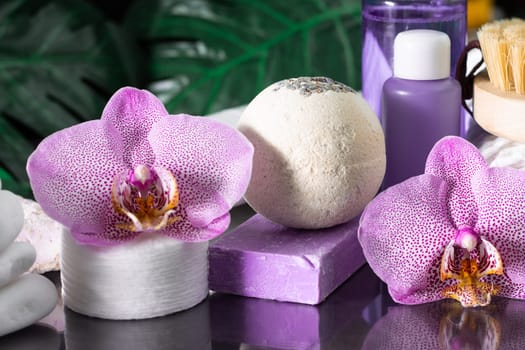 Beautiful lilac orchid flower, cosmetic bottles, cotton pads and handmade soap with stack of white stones and monstera leaves on black surface. SPA or beauty salon and procedures concept