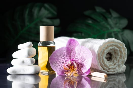 Beautiful lilac orchid flower, clear bottle of yellow oil or perfume, wooden sticks and rolled towel with stack of white stones and monstera leaves, on black surface. SPA salon and procedures concept