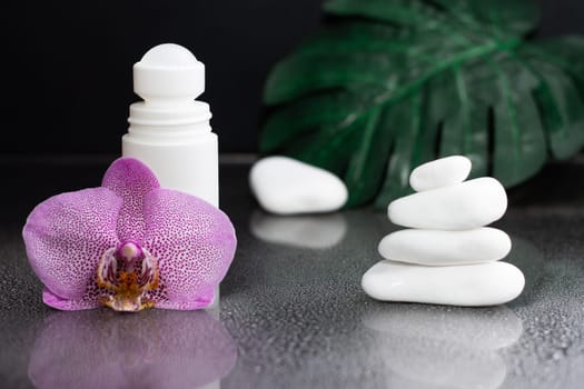 Beautiful lilac orchid flower and white roll-on deodorant with white stones and monstera leaves, on black wet background