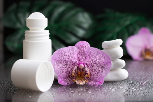 Beautiful lilac orchid flower and white roll-on deodorant with white stones and monstera leaves, on black wet background