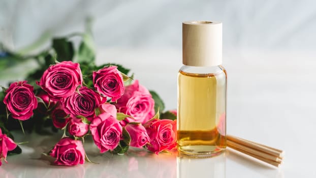 Clear bottle of yellow oil or perfume with wooden sticks and delicate pink roses on white surface. SPA salon and procedures concept. Banner. Copy space