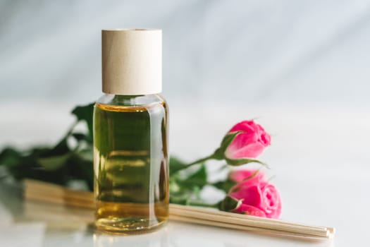 Clear bottle of yellow oil or perfume with wooden sticks and delicate pink roses on white surface. SPA salon and procedures concept. Copy space
