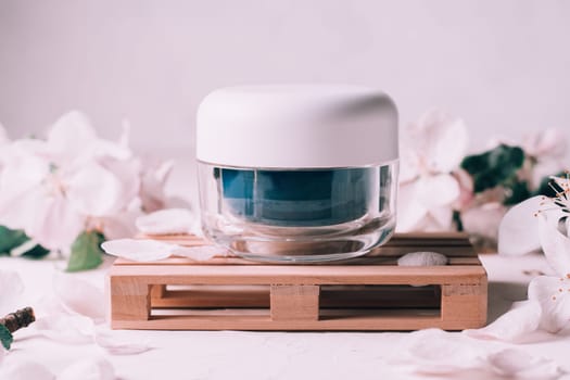 Blue glass jar of cream on wooden podium in form of pallet on light plaster surface with apple flowers
