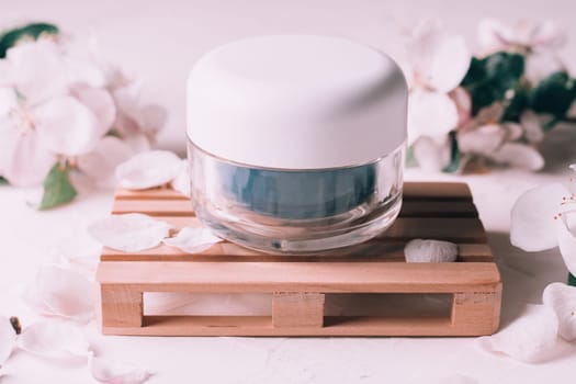 Blue glass jar of cream on wooden podium in form of pallet on light plaster surface with apple flowers