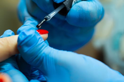 A manicurist in blue gloves, covering the nails with a long-lasting red gel polish.