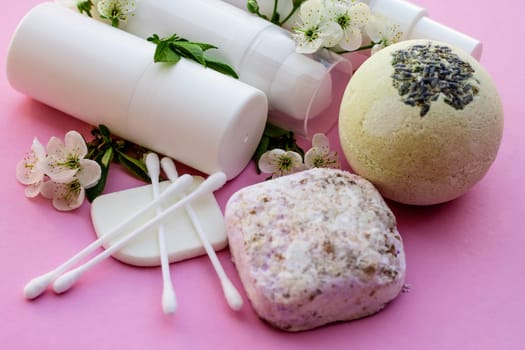 White cosmetic bottles, bath bomb, handmade soap, bath salt, massage brush, sponge, cotton buds with cherry flowers on a pink background. Natural organic cosmetics concept.