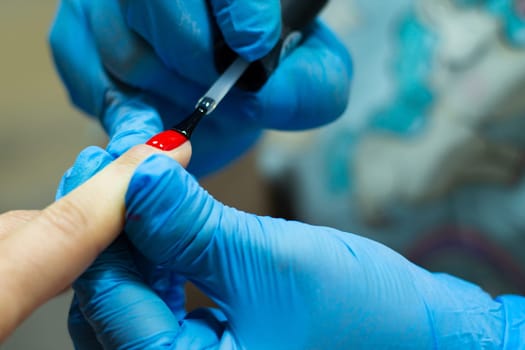 A manicurist in blue gloves, covering the nails with a long-lasting red gel polish.