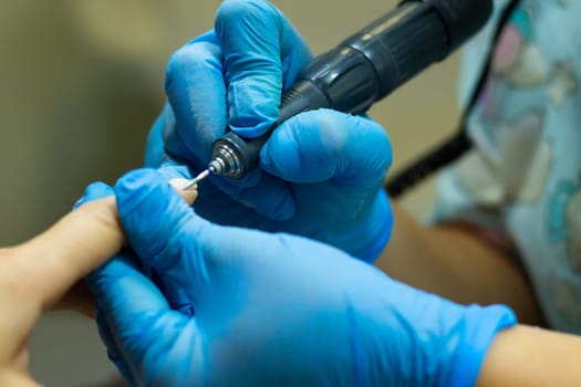 A manicurist in blue gloves makes a manicure with a device, cuticle treatment