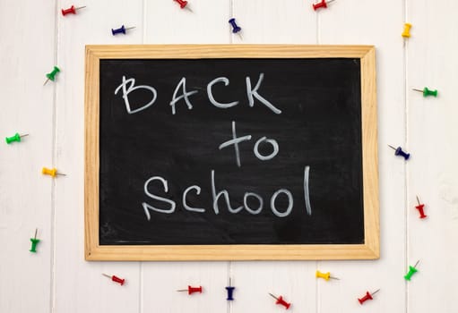Back to school black chalk board on a white wooden background, color pins. Top view. Copy space. Education concept.