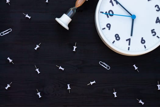 White clock, hourglass and pins on a black wooden background. Top view. Copyspace. Education or business concept.