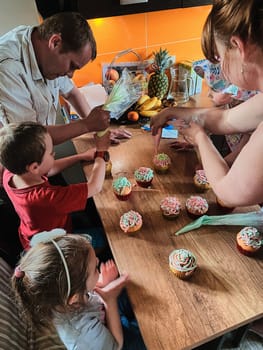 Group of children baking cupcakes, squeezing cream from confectionery bag, preparing ingredients, topping, sprinkles for decorating cookies. Kids cooking, working together in kitchen at home. Concept of happy family