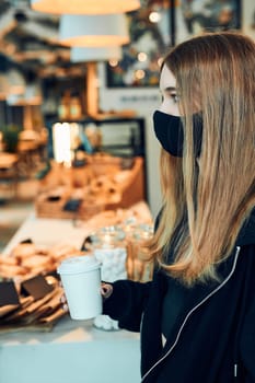 Woman holding coffee cup, standing in queue in a coffee shop and waiting for the order. Person wearing the face mask to avoid virus infection and to prevent the spread of disease in time of coronavirus