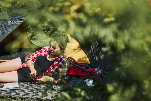 Woman relaxing in tent at camping during summer vacation. Female using smartphone while laying on grass. Actively spending vacations outdoors close to nature. Concept of camp life