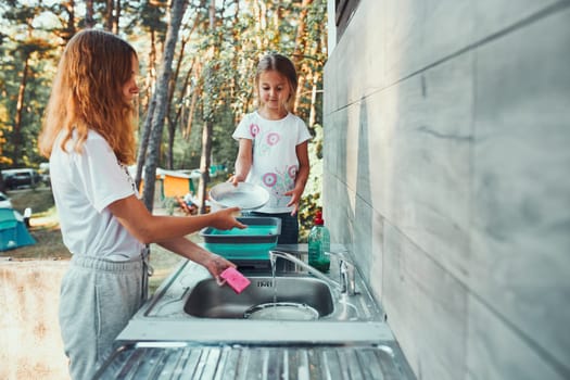Teenager girl washing up the dishes pots and plates with help her younger sister in the outdoor kitchen during vacations on camping. Camp life. Sisters spending time and working together