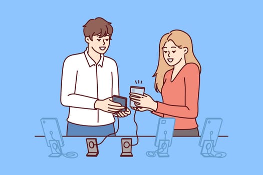 Woman buys new mobile phone in consultation with employee of digital gadget store. Man and girl are standing near showcase with smartphones connected by wire to counter, to avoid stealing phone