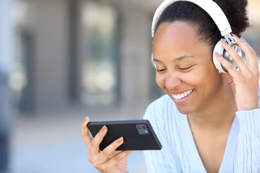 Happy black woman wearing headphone watching videos on horizontal smart phone in the street